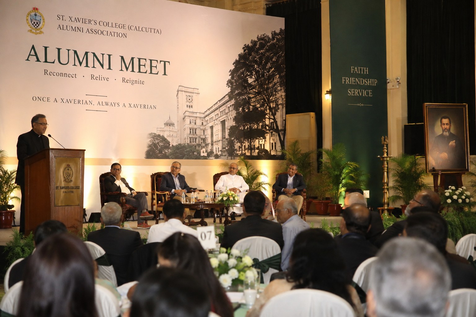 An SXCCAA Alumni Meet in the College hall: a speaker at the lectern before a full house, under a banner reading Reconnect, Relive, Reignite